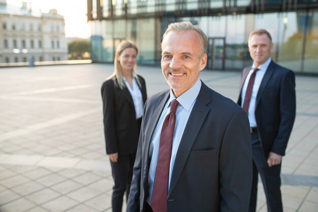 Happy middle aged businessman wearing office suit, standing outdoors and looking at camera. His team standing behind. Teamwork and team success concept