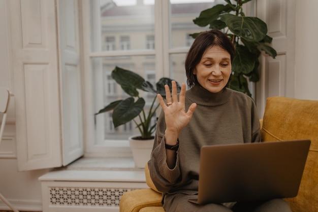 Happy middle aged caucasian woman sitting on sofa and holding laptop while having video call with family friends