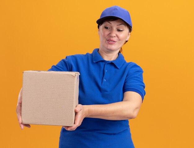 Счастливая женщина-доставщик средних лет в синей форме и кепке держит картонную коробку, глядя на нее, улыбаясь, стоя над оранжевой стеной