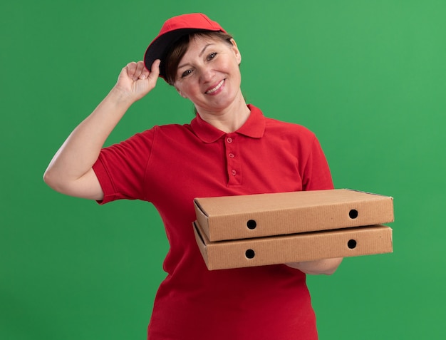 행복 한 중간 세 배달 여자 빨간색 유니폼과 모자 녹색 벽 위에 자신감 서 웃 고 앞에서보고 피자 상자를 들고