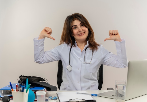 Happy middle-aged female doctor wearing medical robe and stethoscope sitting at desk with medical tools clipboard and laptop pointing at herself isolated