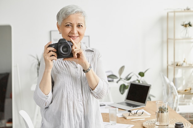 スタイリッシュなオフィスのインテリアでポーズをとって、プロのデジタル一眼レフカメラを保持し、笑顔で短い白髪の幸せな中年女性写真家