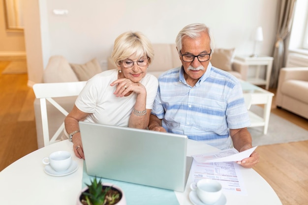 Happy middle aged husband and wife sitting at table with laptop and paper bills calculating domestic incomes together at home