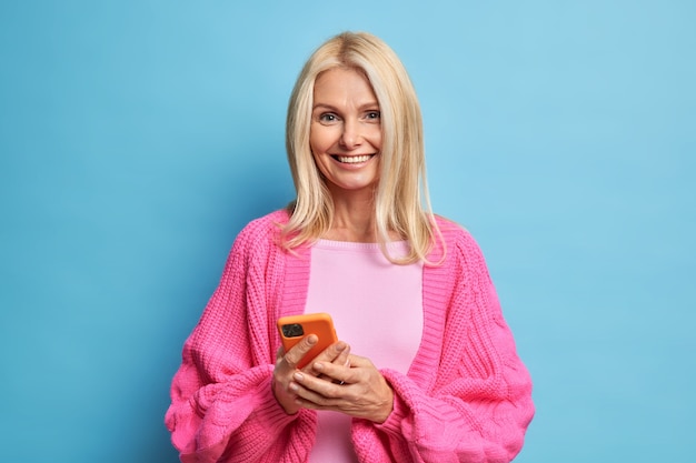 Happy middle aged lady holds mobile phone glad to read pleasant sms chats with friends in social networks wears warm knitted jumper stands indoor. Elderly technology concept. Modern grandmother