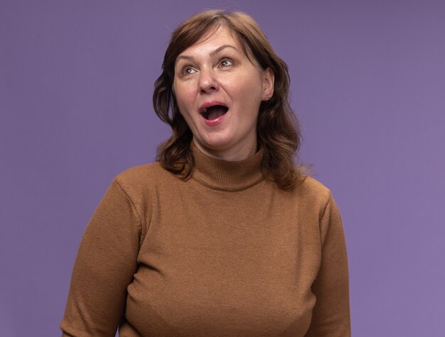 Happy middle aged woman in brown turtleneck looking up happy and cheerful standing over purple wall