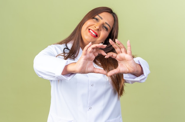 Felice donna di mezza età medico in camice bianco con stetoscopio guardando davanti facendo il gesto del cuore con le dita sorridenti allegramente in piedi sul muro verde