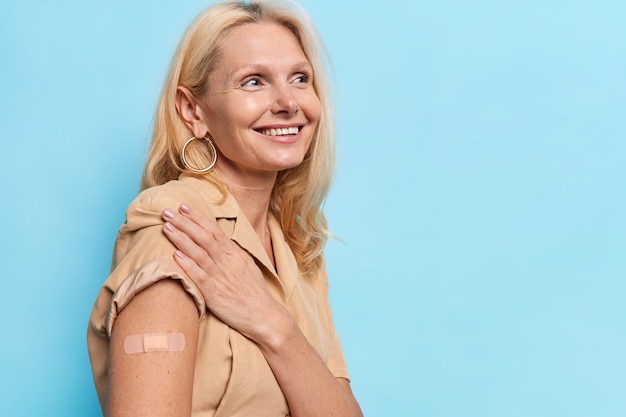 Foto gratuita la donna di mezza età felice ottiene l'inoculazione nella spalla si fa vaccinare in clinica mostra la spalla con il vestito beige della fasciatura adesiva isolato sulla parete blu