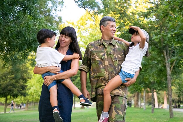 Happy military man walking in park with his wife and kids, teaching daughter to make army salute gesture. Full length, back view. Family reunion or military father concept