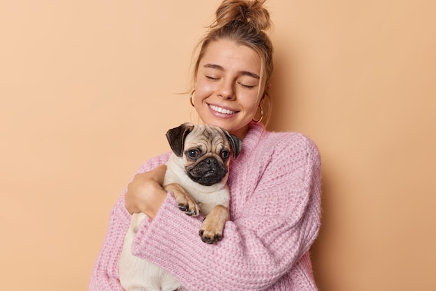 Happy millennial girl with pug dog embraces favorite pet with love keeps eyes closed smiles gently wears knitted sweater isolated over beige background. Good relationships between animals and people