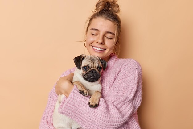 パグ犬と幸せなミレニアル世代の女の子は愛を込めてお気に入りのペットを抱きしめ、目を閉じて笑顔を優しくベージュの背景の上に分離されたニットのセーターを着ています。動物と人との良好な関係