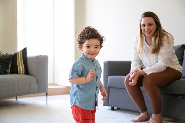 リビングルームに立っている幸せなミックスレースの巻き毛の小さな男の子。ソファに座って笑っている金髪の長髪の母親。セレクティブフォーカス。家族の時間、母性と週末の概念