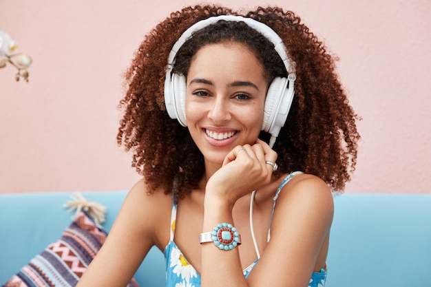 Счастливая женщина-блогер из смешанной расы имеет афро-прическу, слушает любимую радиостанцию в наушниках или наслаждается отдыхом под музыку