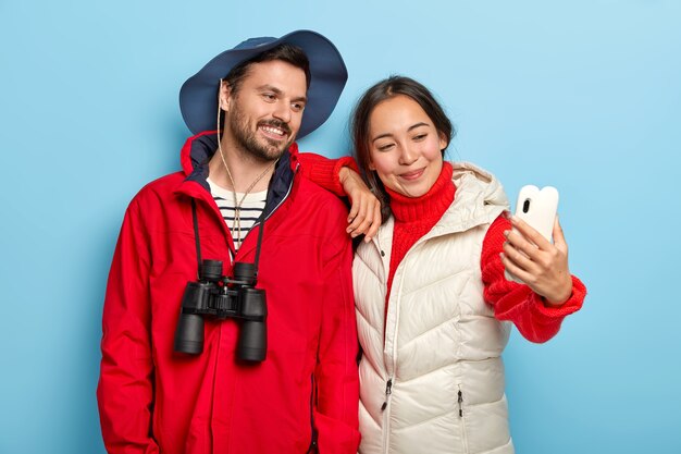 Happy mixed race lovely couple with glad expressions, take selfie on smartphone camera, spend vacation together, dressed casually, use binoculars