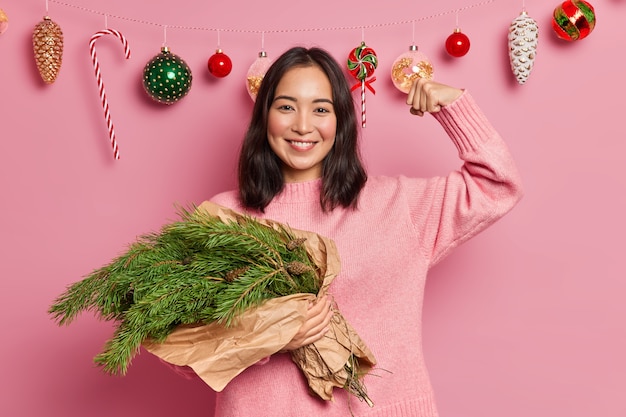 Happy mixed race woman holds bouquet of fir tree branches raises arm and shows how strong she is dressed in sweater poses 