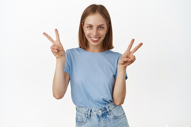 幸せな現代のブロンドの女の子は、平和のvサインを示し、カワイイ笑顔で、白い背景の上に夏のTシャツに立って、積極性、楽しさと喜びを表現します。
