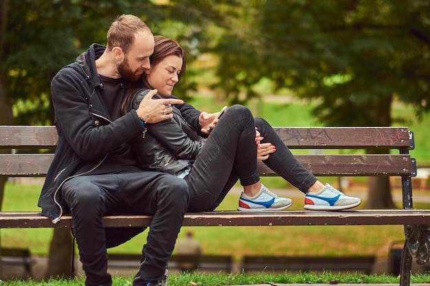 公園のベンチに寄り添いながらインターネットで何か楽しいものを閲覧している幸せな現代のカップル。彼らの愛と自然を楽しんでいます。