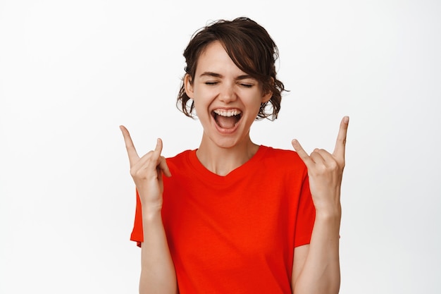 Felice ragazza moderna che fa festa, mostrando lingua e corna di metallo pesante, gesto rock n roll, sorridendo e godendosi il divertimento del concerto su bianco