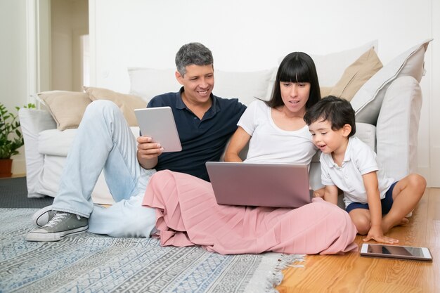 Happy mom, dad and cute little boy using computers, sitting on apartment floor, enjoying leisure time together.