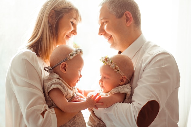 Happy mom and dad pose with funny twins on their arms before a bright window 