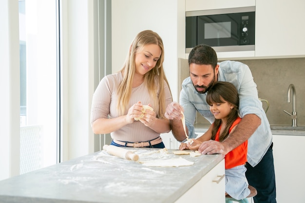 Happy mom and dad teaching daughter to knead dough at kitchen table with flour powder.