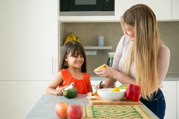 Счастливая мама и дочь готовят салат с лимонной заправкой. Девочка и ее мать чистят и режут овощи на кухонном столе, болтают и веселятся. Семейная кулинария или концепция здорового питания