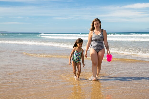 幸せなママと小さな女の子は水着を着て、ビーチで海の水に足首を深く歩いています