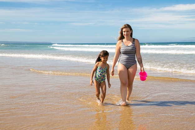 Happy mom and little girl wearing swimsuits, walking ankle deep in sea water on beach