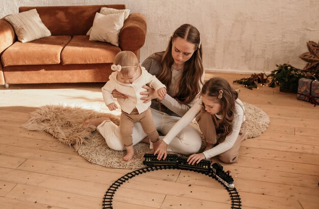 Счастливая мама играет с маленькими дочками с игрушечной железной дорогой на полу в комнате