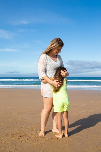 Happy mom and sweet little daughter in summer clothes hugging while standing on ocean beach