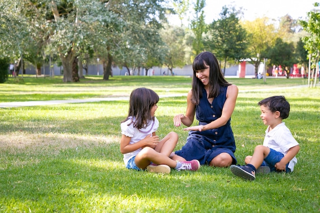 Happy mom and two kids sitting on grass in park and playing. Cheerful mother and children enjoying leisure time in summer. Family outdoors concept