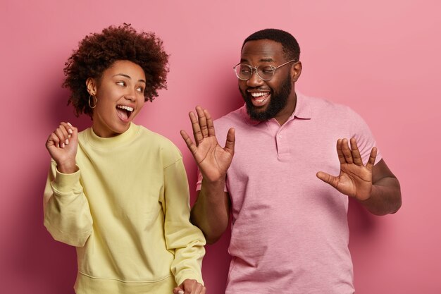 Happy mood and entertainmet. Positive good humored Afro American couple dance with arms raised, listen music, sing along, move actively