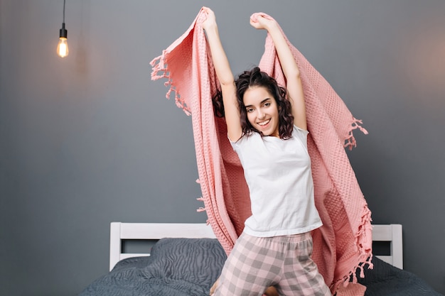 ピンクの毛布が付いているベッドでストレッチうれしそうな若い女性の近代的なアパートで自宅で幸せな朝。笑顔で真のポジティブな感情を表現