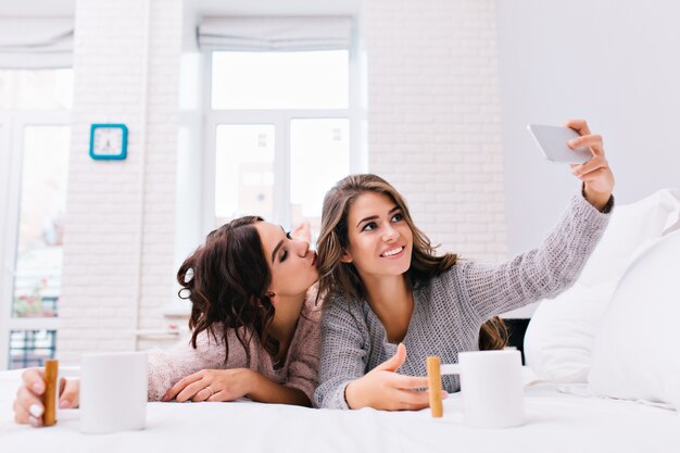 Happy morning of two joyful attractive girls making selfie on white bed. Pretty young women having fun together, smiling, chilling, drinking coffee,  friends .