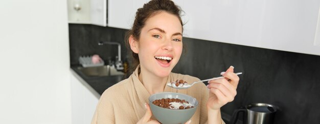 Free photo happy mornings gorgeous young woman eating cereals with milk standing in kitchen with breakfast bowl