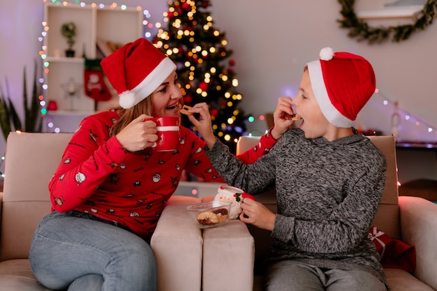 無料写真 幸せな母とサンタの帽子をかぶった小さな息子とお茶のカップを背景にクリスマスツリーと飾られた部屋で楽しんでソファに座ってクッキーを食べる