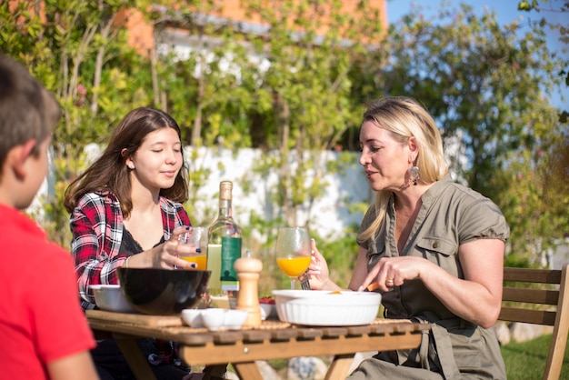 幸せな母と子供たちは裏庭でバーベキューを食べています。中年の大人の女性と子供たちがテーブルの周りに座って、作りたての食べ物を食べ、飲み物を飲みます。バーベキュー、料理、食べ物、家族のコンセプト