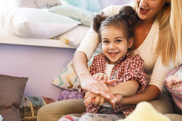 Happy mother and daughter are playing in room near window dressed in casual clothes