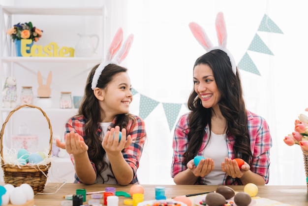 Happy mother and daughter holding easter eggs on hand looking at each other at home