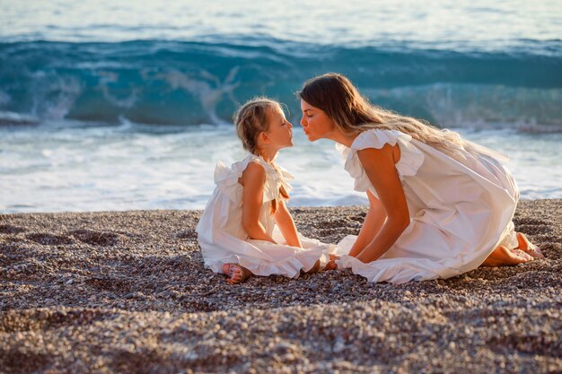 Happy mother and daughter sitting together and kissing each other in seashore in white dress during sunset .