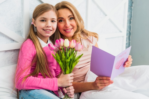 Madre felice e figlia che si siedono con i fiori e la cartolina d'auguri