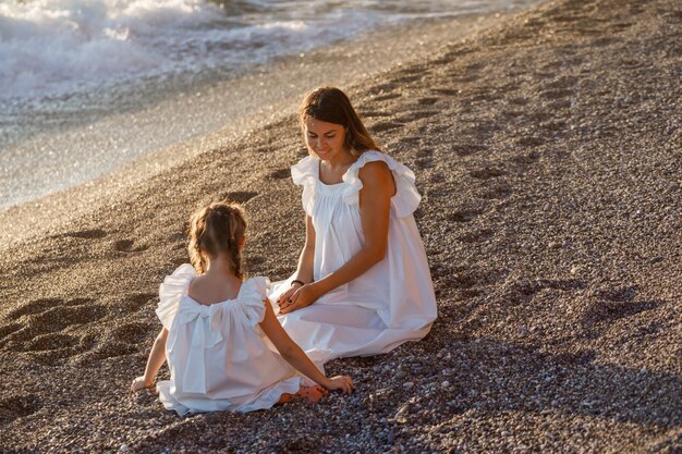 Счастливая мать и дочь в белом платье, сидя на песке на берегу моря во время заката