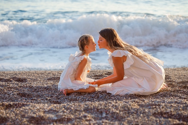 Happy mother and daughter in white dress sitting together and kissing each other in seashore during sunset .