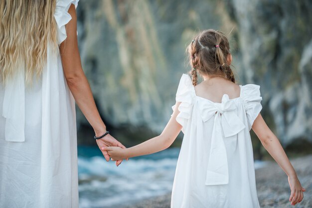 Happy mother and daughter in white dress standing and holding hands in seashore during sunset .