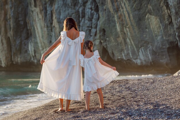 Счастливая мать и дочь в белом платье стоя и держа их платья на берегу моря во время заката. вид сзади