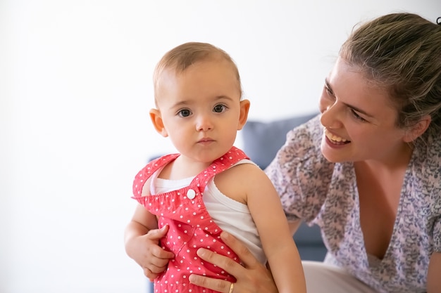 幸せな母親は女の赤ちゃんを抱いて、笑顔で彼女を見ています。赤いダンガリーショーツの深刻な愛らしい幼児。子供と話しているかわいいお母さん。家族の時間と母性の概念