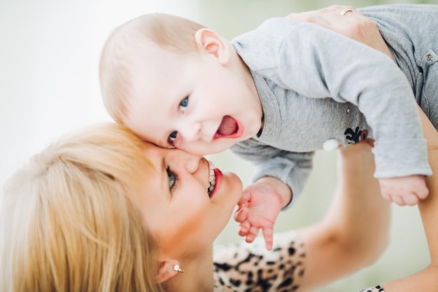 Happy mother holding loving her baby and toching by noses, looking each other. Blondie pretty mum with little kid having fun, playing and spending time together. Concept of family and love.