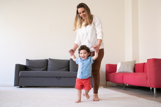Happy mother holding son hands and teaching him to walk. Cheerful curly mixed-race little boy walking on carpet barefoot with help of long-haired mom. Family time, childhood and first step concept