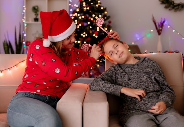 壁​に​クリスマスツリー​が​飾られた​部屋​の​ソファ​に​座っている​彼女​の​眠っている​息子​の​頭​に​面白い​縁​を​置く​サンタ​帽子​の​幸せな​母親