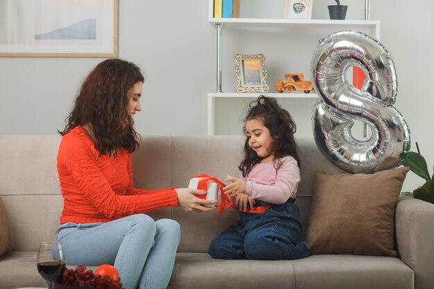 Счастливая мать получает подарок от своей маленькой дочери, сидящей на диване с поздравительной открыткой, весело улыбаясь в светлой гостиной, празднование Международного женского дня 8 марта
