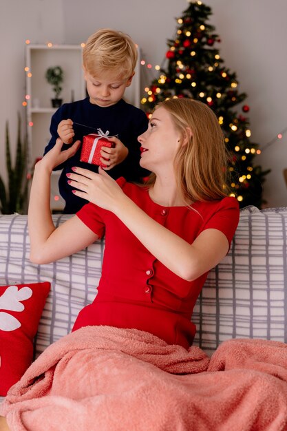 Happy mother in red dress sitting on a couch smiling  while her little child standing behind giving a gift to his   mother in a decorated room with christmas tree in the wall